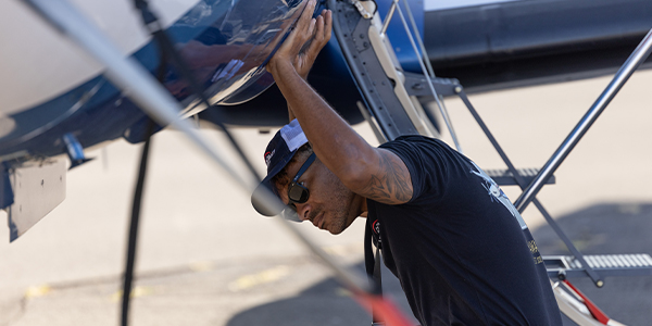 Aircraft mechanic wearing a black shirt, sunglasses, and a baseball cap works beneath a helicopter, reaching upward to inspect or adjust a component on the aircraft’s underside on a paved surface.
