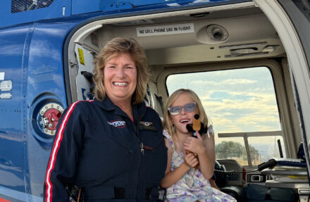 An adult stands at the open side door of a blue medical helicopter alongside a child seated just inside the doorway. The helicopter interior, door frame, and exterior fuselage are visible, with a safety sign above the doorway reading “NO CELL PHONE USE IN FLIGHT,” and daylight visible through the opposite windows.