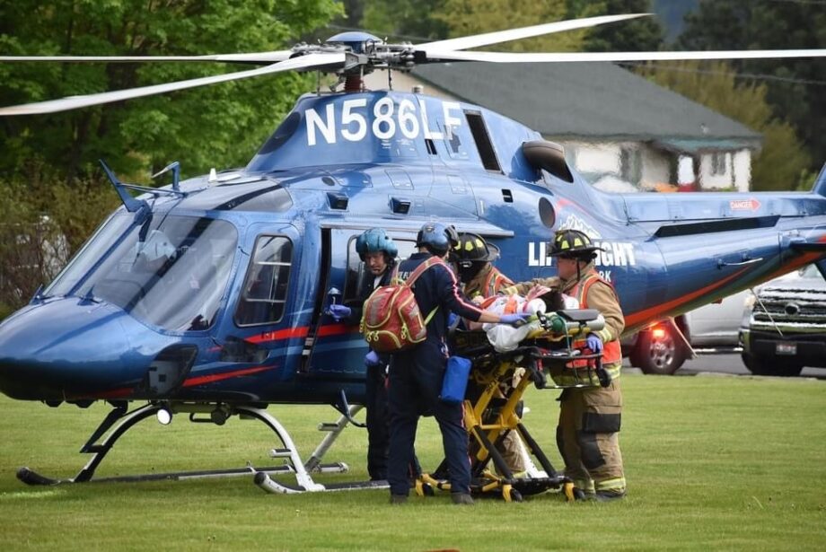 A medical team transfers a patient on a wheeled stretcher into a blue Life Flight Network helicopter parked on a grassy landing area. Several responders stand around the open cabin door guiding the stretcher, while the helicopter’s registration number N586LF, landing skids, windows, and medical transport branding are visible. Trees, vehicles, and nearby buildings appear in the background.