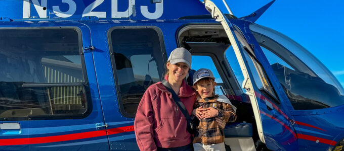 An adult and a young child stand beside an open blue helicopter door on a sunny day. The helicopter’s exterior, windows, and landing area are visible in the background.