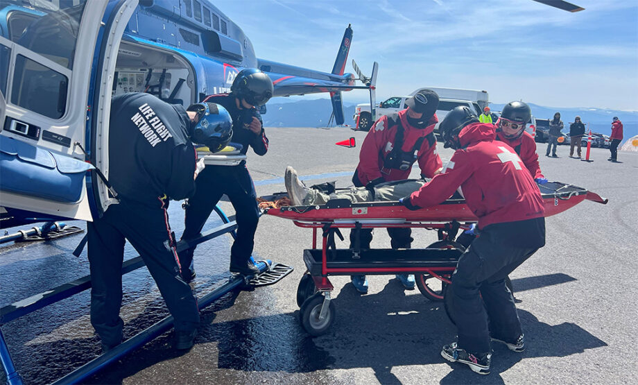 Several medical responders work together to transfer a patient on a wheeled stretcher into a blue Life Flight Network helicopter parked on a paved landing area. The helicopter’s side door is open, crew members wear helmets and protective gear, and the aircraft’s skids, interior, and medical transport branding are visible. Other responders and cones appear in the background under a clear blue sky.