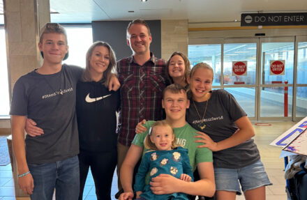 Seven people of varying ages pose together indoors near sliding glass doors at a building entrance. Visible signage on the doors reads “Do Not Enter,” and the group stands closely with arms around one another, while one child sits in front. The setting appears well lit with tiled floors and posted informational signs nearby.