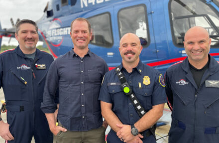 Four adults stand shoulder to shoulder in front of a blue Life Flight Network helicopter on an airfield. The helicopter’s windows, rotor mast, and medical transport branding are visible in the background, and several of the individuals wear flight or emergency services uniforms.