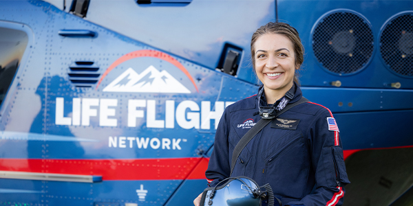 Person wearing a dark blue flight uniform holds a helicopter helmet while standing in front of a blue medical helicopter with ‘Life Flight Network’ text and mountain logo visible on the aircraft’s side.