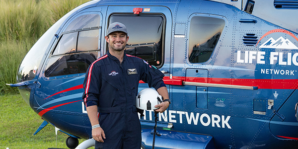 Person in a dark blue flight suit holding a white helicopter helmet while standing beside a blue medical helicopter with ‘Life Flight Network’ text on the side; grassy field and daylight setting.
