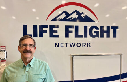 An adult stands indoors in front of a white wall displaying the “Life Flight Network” logo with a mountain graphic. Part of a metal railing or equipment frame is visible below the logo, and wall-mounted fixtures appear to the left.