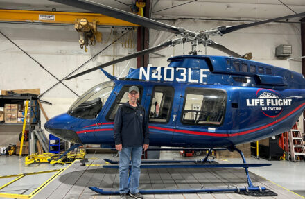 An adult stands inside an aircraft maintenance hangar in front of a blue Life Flight Network helicopter with registration markings visible on the tail boom. The helicopter rests on its skids with rotor blades overhead, and tools, equipment, and storage shelves are visible in the background of the hangar.