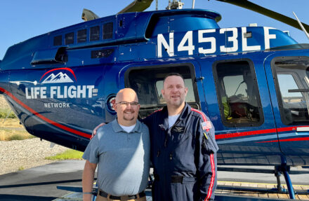 Two adults stand on a paved helipad in front of a blue Life Flight Network helicopter with registration markings visible on the fuselage. The helicopter’s side door, windows, landing skids, and medical transport branding are clearly visible in bright daylight.