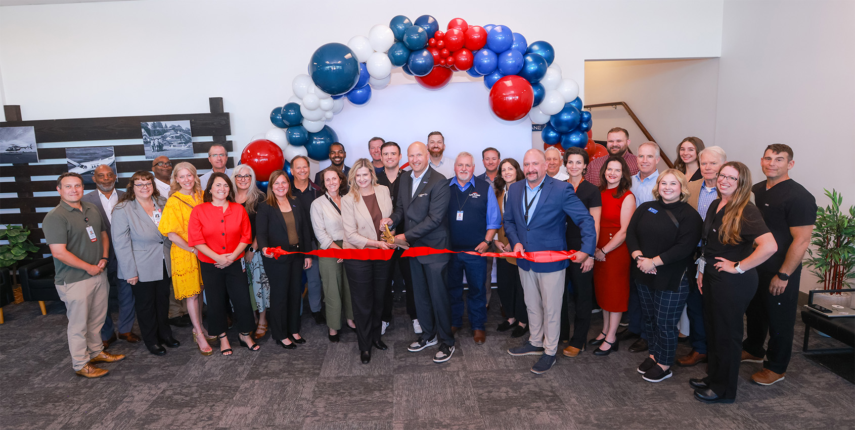 Group photo of staff and community members gathered for a ribbon-cutting ceremony beneath a red, white, and blue balloon arch.
