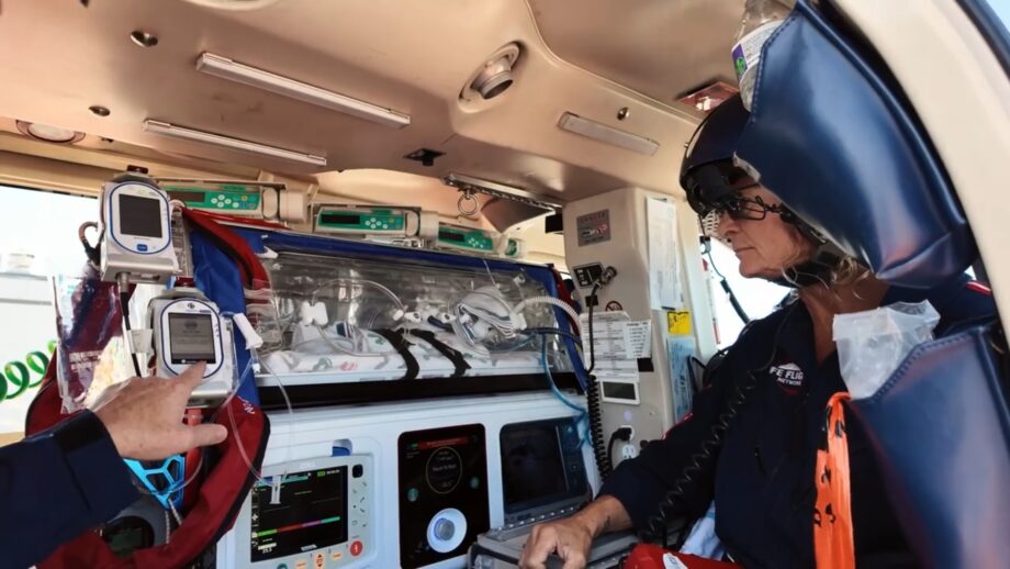 Inside a medical helicopter, a transparent transport incubator is secured to a stretcher and connected to multiple monitors, infusion pumps, and medical tubing. A crew member wearing a headset sits beside the equipment, while another hand gestures toward a device. The helicopter cabin ceiling, control panels, and mounted medical instruments are clearly visible.