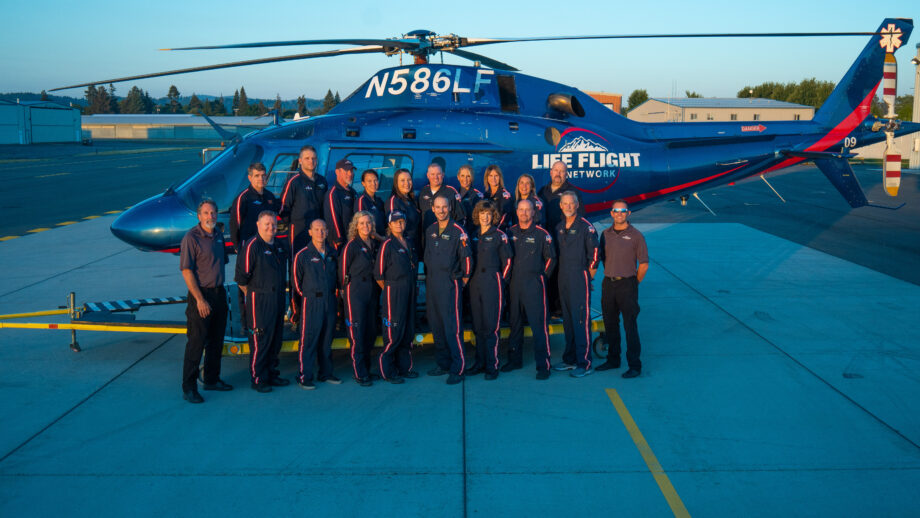 A group of 21 people, the Life Flight neonatal pediatric team, pose in front of a helicopter. The sun is setting in the background.