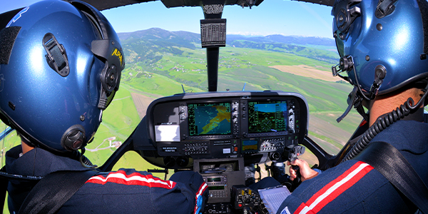 View from inside a small aircraft cockpit showing two people wearing blue flight helmets seated side by side, hands on the controls, with digital flight displays in front of them and green farmland, hills, and mountains visible through the windshield below.