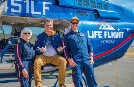 Three people stand and sit at the open side door of a blue medical helicopter marked “Life Flight Network” on an airport tarmac. Medical equipment is visible inside the aircraft, and the helicopter’s exterior, rotor housing, and landing surface are clearly shown in bright daylight.