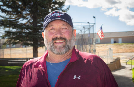 David Swanson wears a blue Life Flight ballcap. He has a salt-and-pepper beard, a wears burgundy athletic shirt over a medium blue undershirt. A baseball backstop, green grass, and fir tree are behind him.
