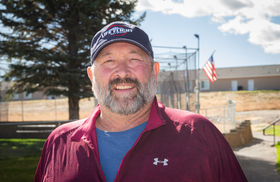 David Swanson wears a blue Life Flight ballcap. He has a salt-and-pepper beard, a wears burgundy athletic shirt over a medium blue undershirt. A baseball backstop, green grass, and fir tree are behind him.