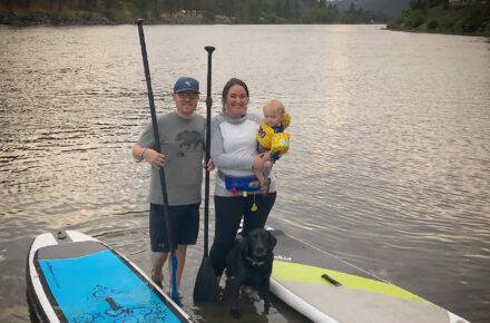 Desirae and her partner stand in shallow water, next to stand-up paddle boards. They have paddles in hand, and she a young baby on her arm.