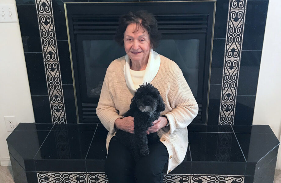 Older woman sits in front of fireplace with a small, curly-haired dog sitting on her lap.