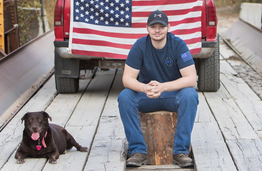 A man sits on a cut log with a brown lab dog laying next to him. A red truck with an American flag draped over the tailgate is parked behind him.