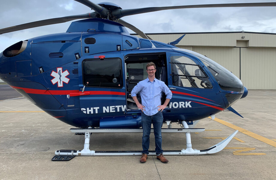 Young man in light blue shirt and blue jeans, stands with hands on hips, posing in front of a Life Flight Network helicopter