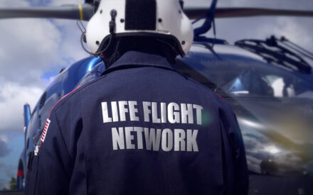 Pilot seen from behind with helicopter in background. "Life Flight Network" emblazoned on back of jumpsuit.