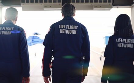 Three crew members are seen from behind, walking toward a light-drenched landing pad "Life Flight Network" is emblazoned on the backs of their jumpsuits.