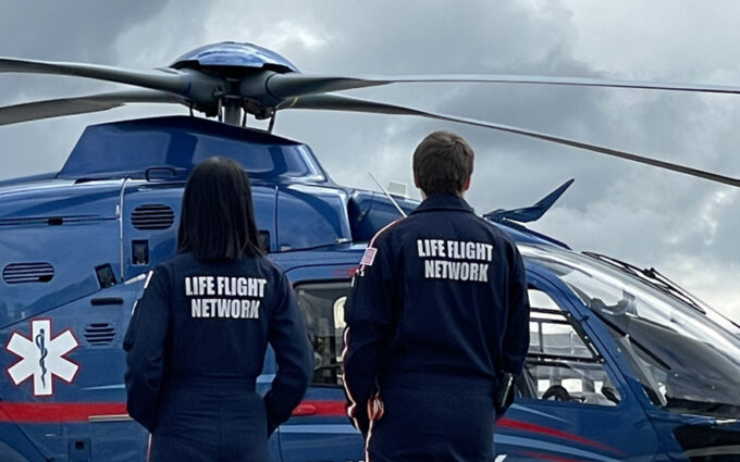 Two people stand in front of helicopter with back facing camera, wearing blue and red jump suites with "Life Flight Network" written across the back.