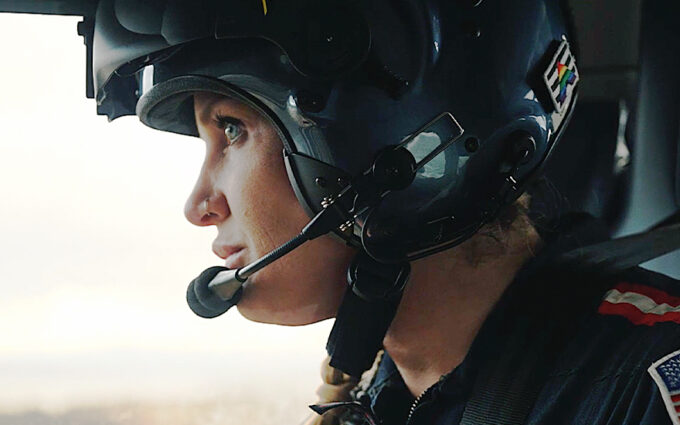 Close up of female flight nurse looks out window of a helicopter. She has blue eyes, a nose ring and is wearing a flight helmet with her hair in a braid.