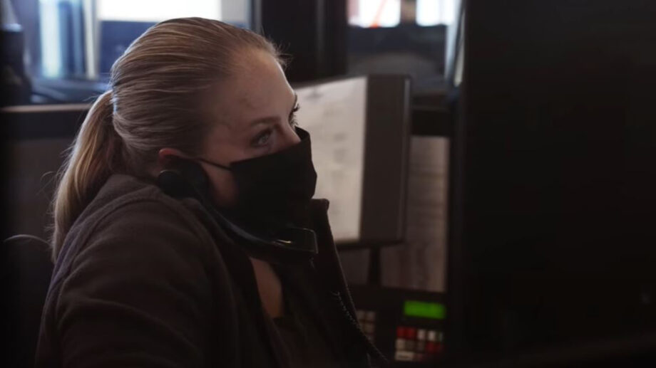 Woman in black medical face mask speaks on phone while looking at a computer screen