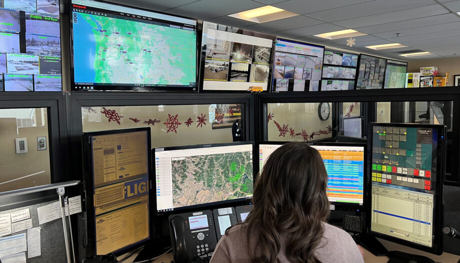 Wide view of communications center. TV screen span length of wall showing vital comms info. Room is sectioned off with cubicles. Staff are answering dispatch request and directing communications.