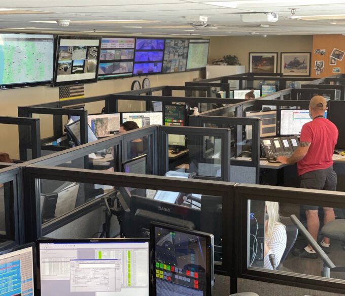 Wide view of communications center. TV screen span length of wall showing vital comms info. Room is sectioned off with cubicles. Multiple staff are answer dispatch request and directing communications.