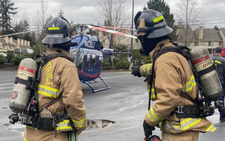 Two fire fighters are seen from behind, pointing at helicopter landing in a parking lot