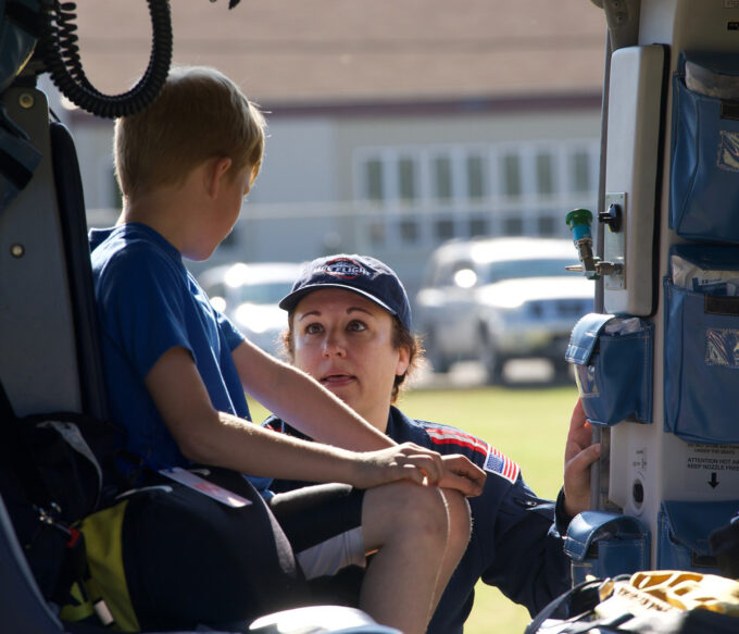 Young boy sits un jump seat of helicopter. Flight nurse gives him tour of the aircraft.