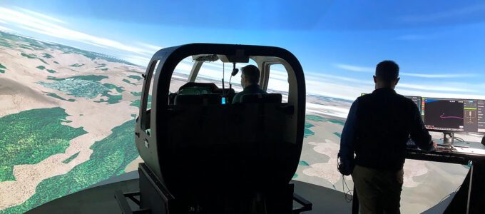 Pilot inside of flight simulator. A skeleton cockpit of a helicopter is seen with a large, wrap around screen showing a flight simulation. A man stands off to side observing a computer screen.