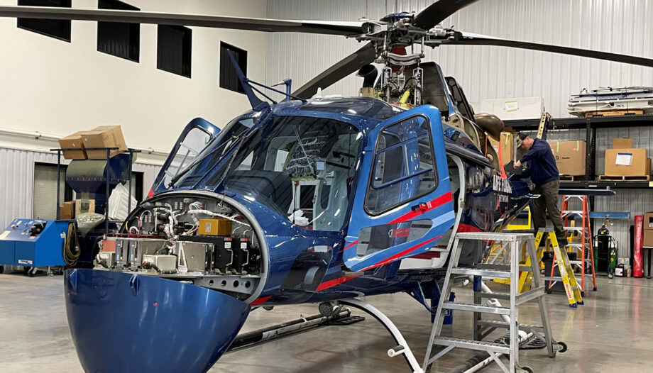 Helicopter in maintenance bay with front nose open, man standing on ladder working on aircraft