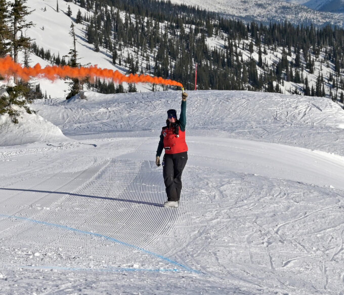 Member of rescue team stands atop snowing mountain holding a smoking flare indicating landing zone