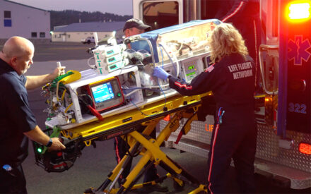 Three people load neonatal transportation chamber into the back of a ground ambulance