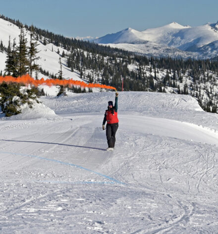 Member of rescue team stands atop snowing mountain holding a smoking flare indicating landing zone