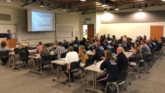 Large group of people in classroom setting. A man at a podium gives a presentation with aid of a large screen.