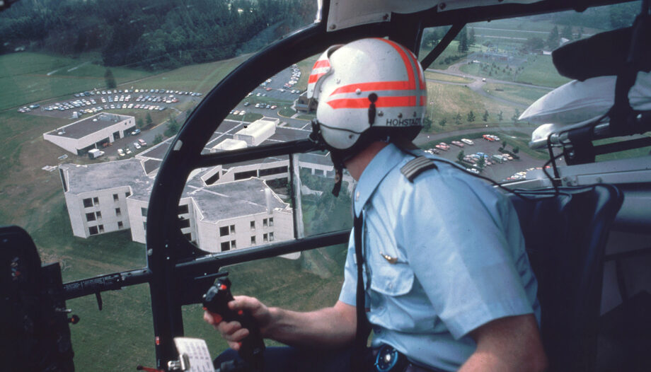 A historical photo from the '70s shows a man piloting a helicopter to a landing zone. Emanuel Hospital can be see out of the cockpit window, over his right shoulder. he appears to be turning at a steep angle.